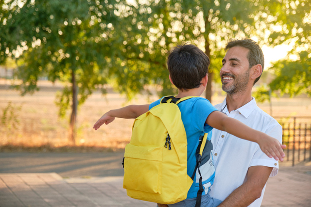 smiling dark-haired father in white shirt holding child in arms in blue t-shirt and yellow backpack
